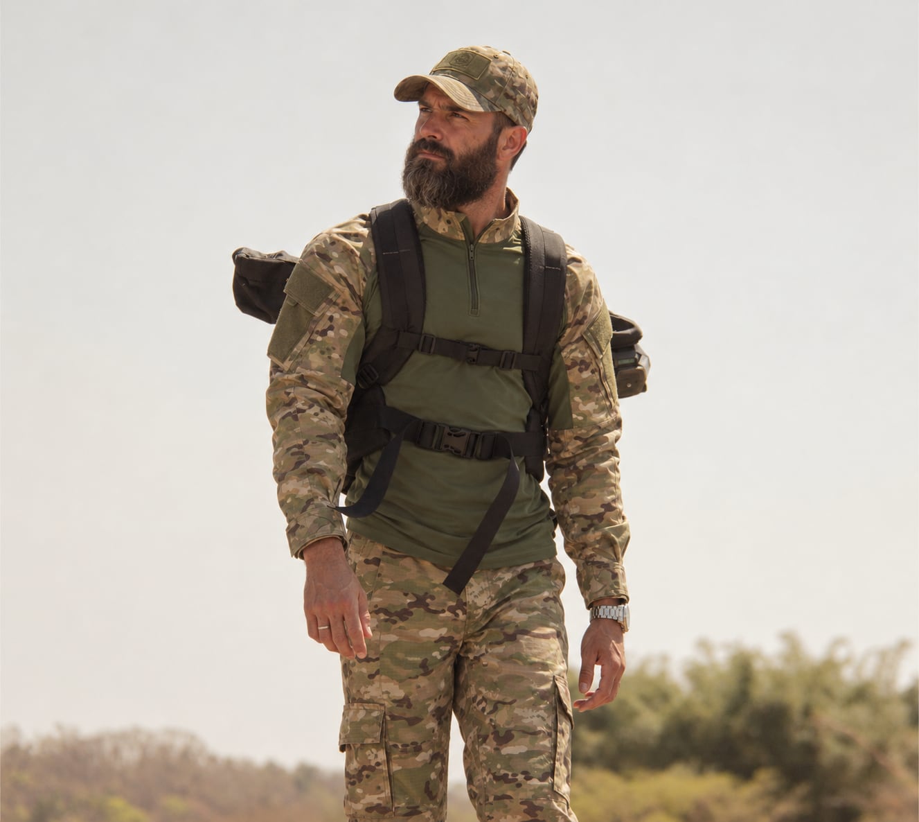 Homem de barba em campo com uniforme camuflado, boné e mochila Fox Boy